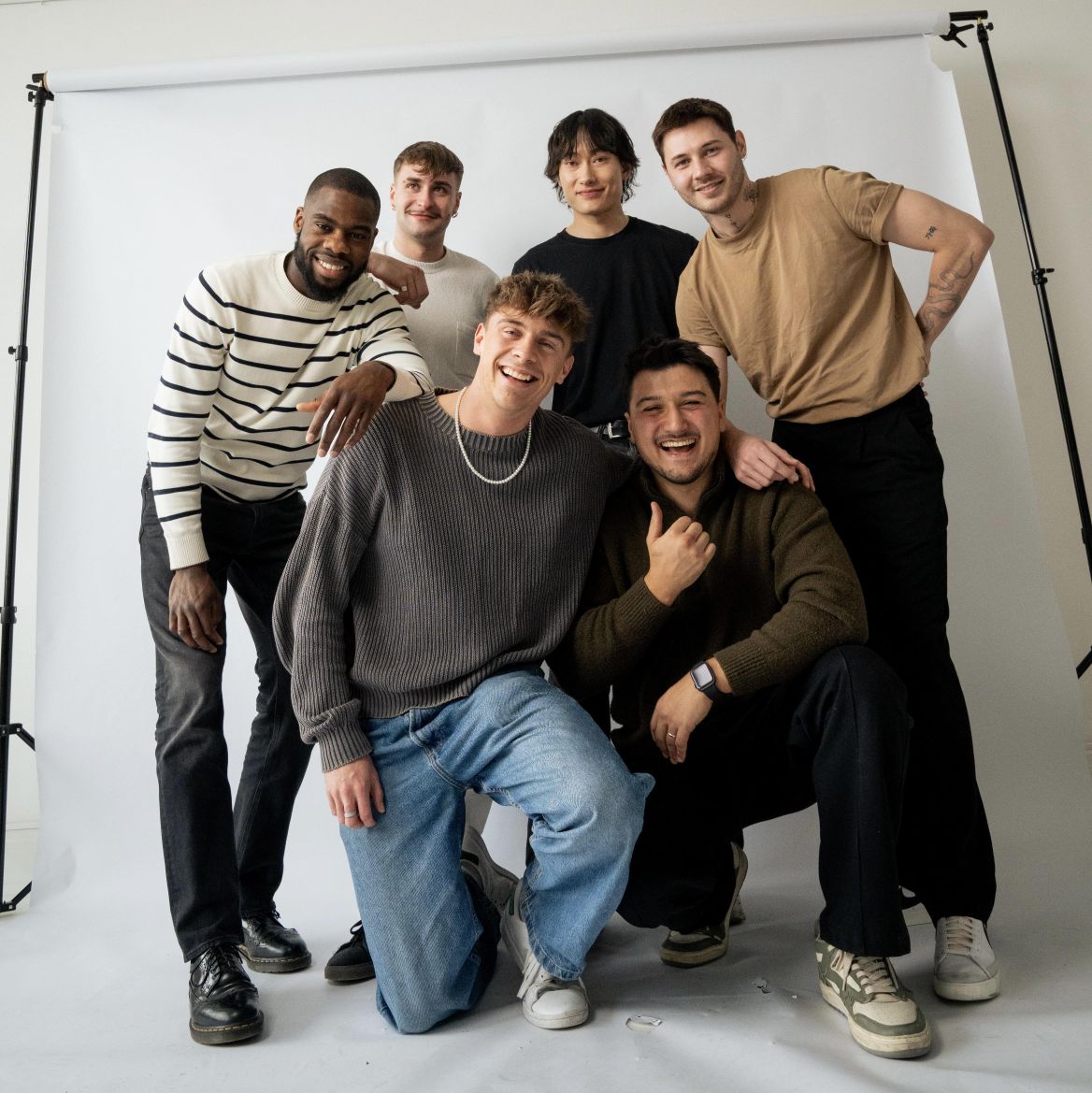 Gruppe junger, stilvoll gekleideter Männer posiert fröhlich vor einem weißen Fotohintergrund – modernes Teamfoto mit entspannter Atmosphäre